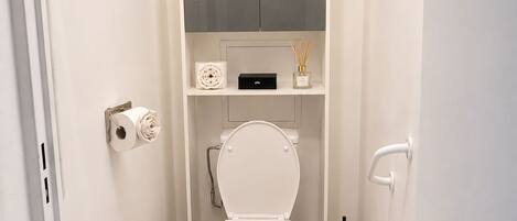 A small, private bathroom with a toilet, a toilet paper holder, a trash bin, a bidet, and a cabinet above the toilet.