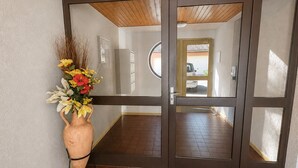 A hallway with a large potted plant, a wooden ceiling, and a view into another room through a glass door. - The Butterfly Garden (La Buisse)