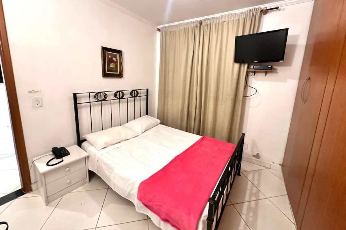 A bedroom with a bed, a nightstand, a TV mounted on the wall, and a window with curtains.