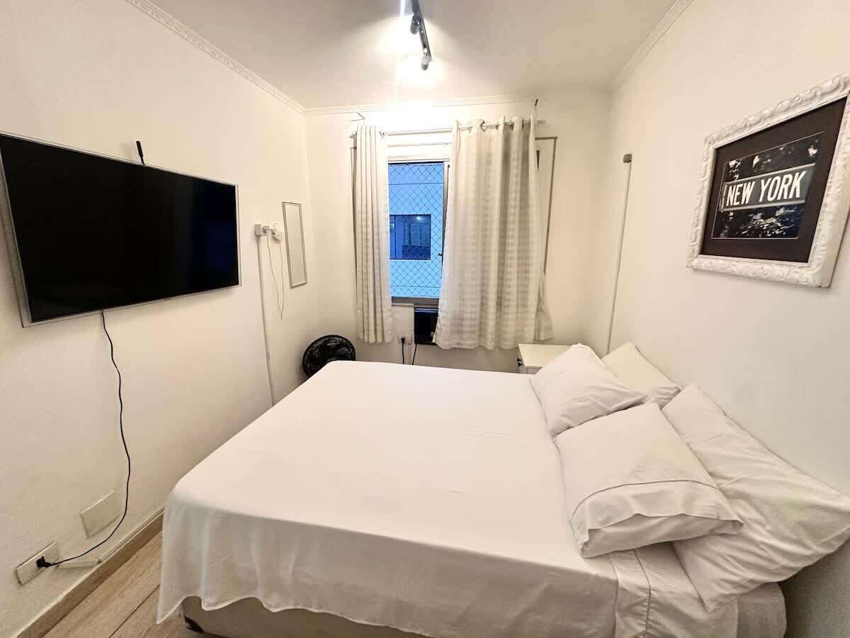 Ein Hotelzimmer mit Bett, Wandfernseher, Fenster mit Vorhängen und einem gerahmten Bild an der Wand.