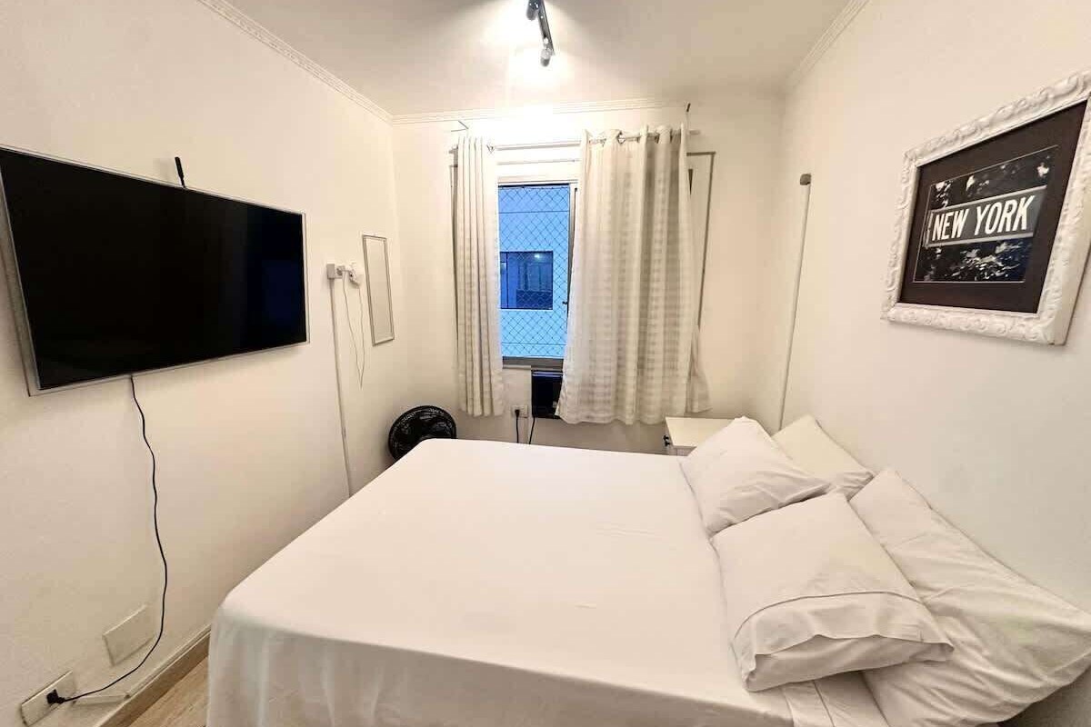 Ein Hotelzimmer mit Bett, Wandfernseher, Fenster mit Vorhängen und einem gerahmten Bild an der Wand.
