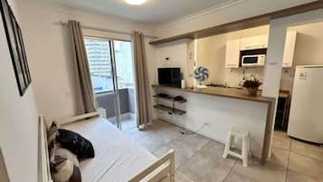 Quarto compacto com cama, pequeno espaço de cozinha, televisão e janela com cortinas.