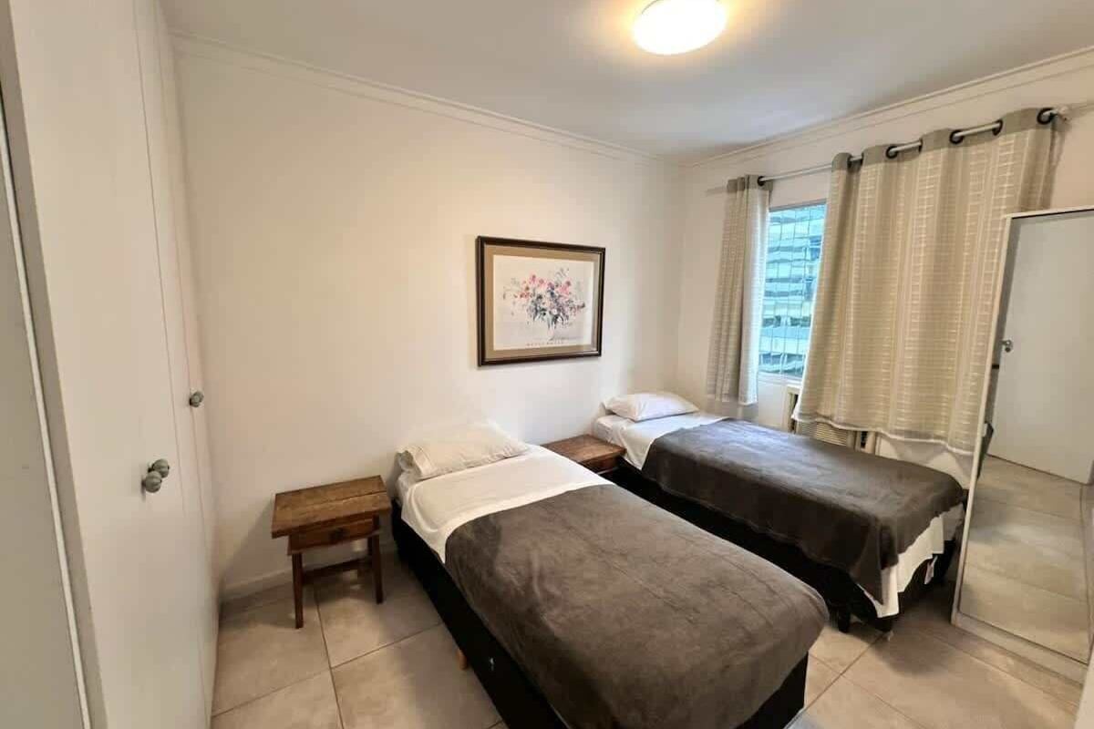 Ein Zimmer mit zwei Betten, einem Nachttisch, einem gerahmten Bild an der Wand und einem Fenster mit Vorhängen.