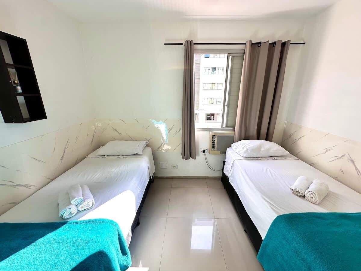A hotel room with two beds, a window with curtains, a wall-mounted TV, and a small shelf.