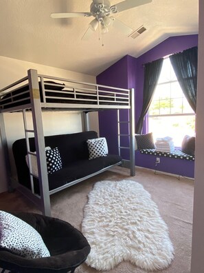 A bunk bed with a white ceiling fan, a black sofa with polka dot pillows, a white furry rug, and a window with curtains.