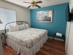 A bedroom with a bed, a ceiling fan, a window with blinds, a painting, and a TV.