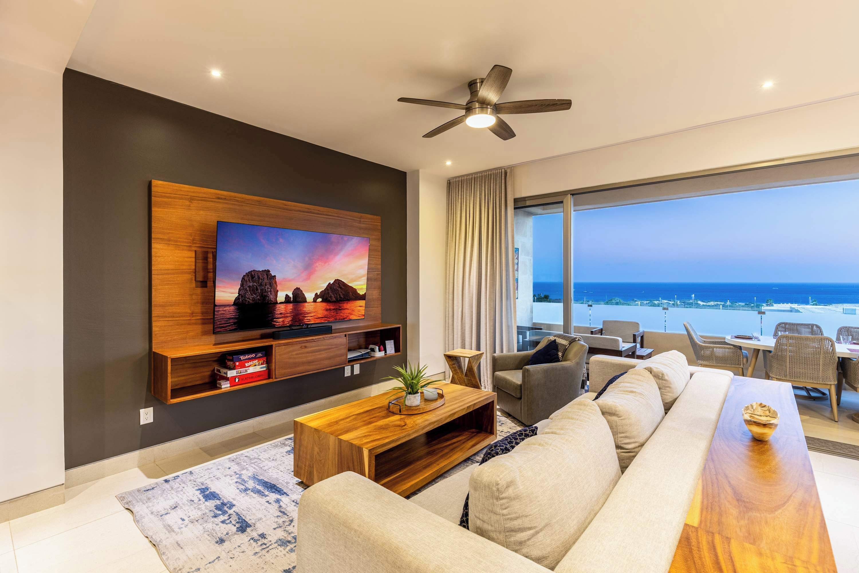 A modern living room with a large TV, a ceiling fan, and a view of the ocean.
