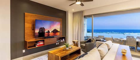 A modern living room with a large TV, a ceiling fan, and a view of the ocean.