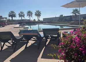 A poolside area with lounge chairs and an umbrella, surrounded by palm trees and flowers. - Two Condos | Ideal for Families & Groups (Cabo San Lucas)