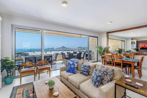 A spacious living area with a large window overlooking a cityscape, a dining area with a view, and a kitchen with a large mirror. - Two Condos | Ideal for Families & Groups (Cabo San Lucas)