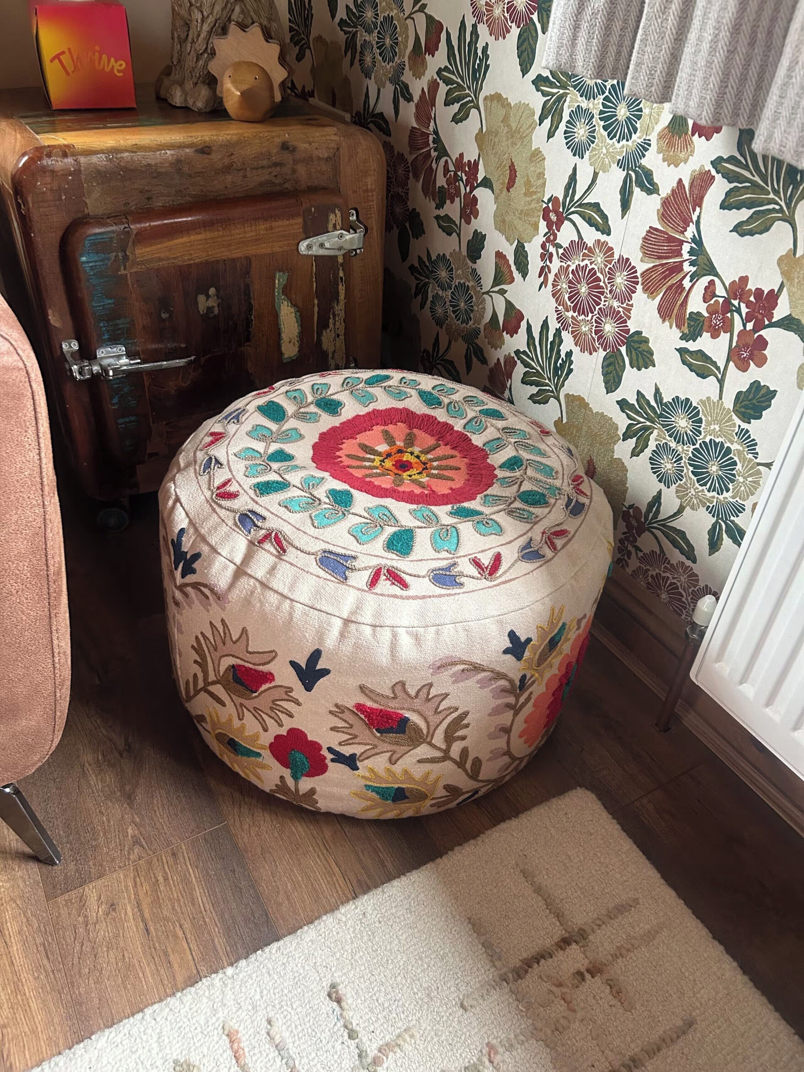 A round ottoman with floral embroidery, a wooden chest, and a patterned wall.