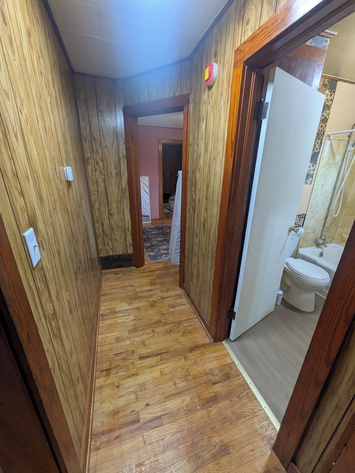 A narrow hallway with wooden walls and a wooden floor, leading to a bathroom with a toilet and a bathtub.