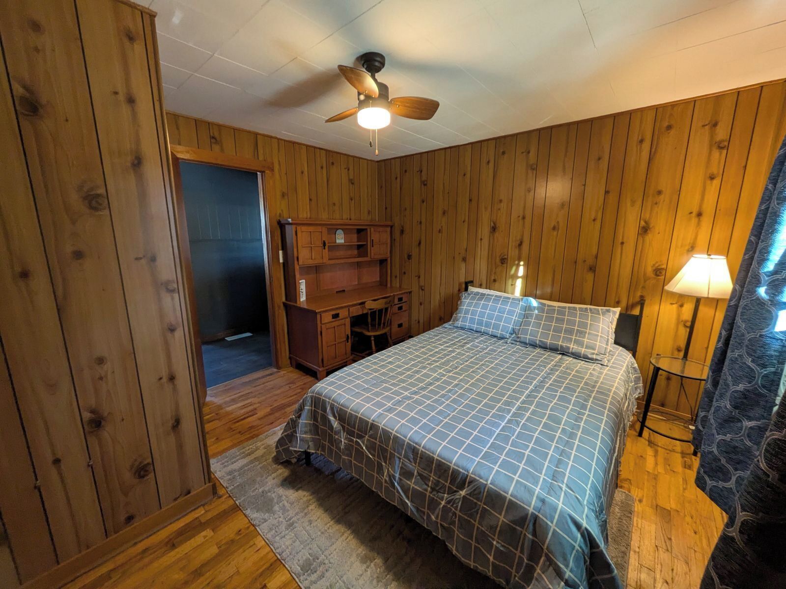 Een slaapkamer met houten lambrisering, een bed met een geruit sprei, een bureau, een stoel, een plafondventilator en een nachtkastje met een lamp.