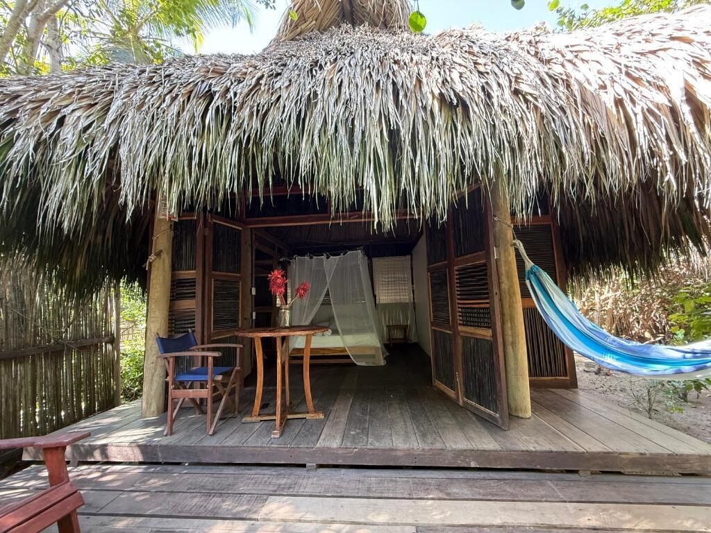 A thatched-roof hut with a wooden deck, a table with chairs, and a hammock.
