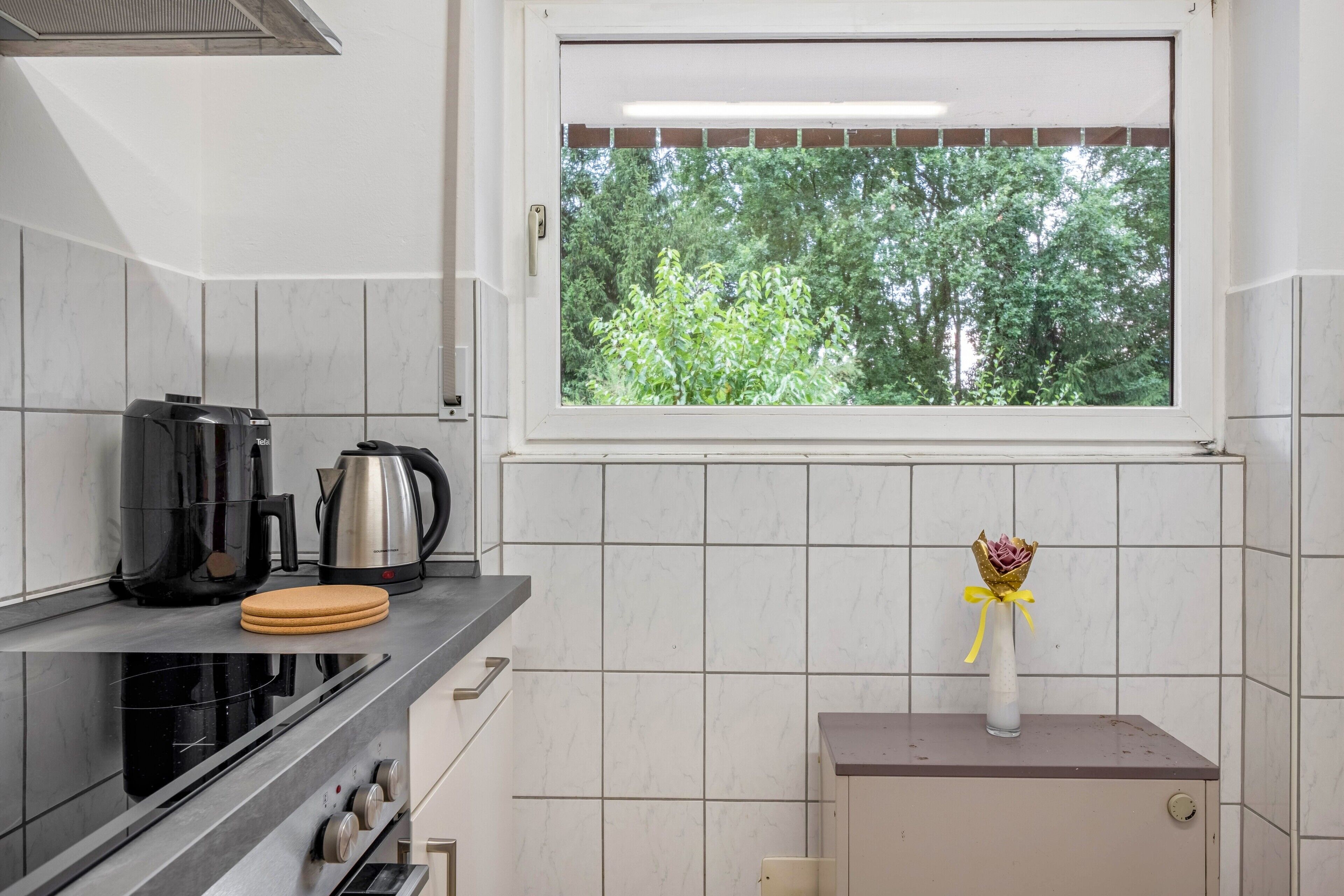A kitchen with a window, a stove, a kettle, and a small cabinet.