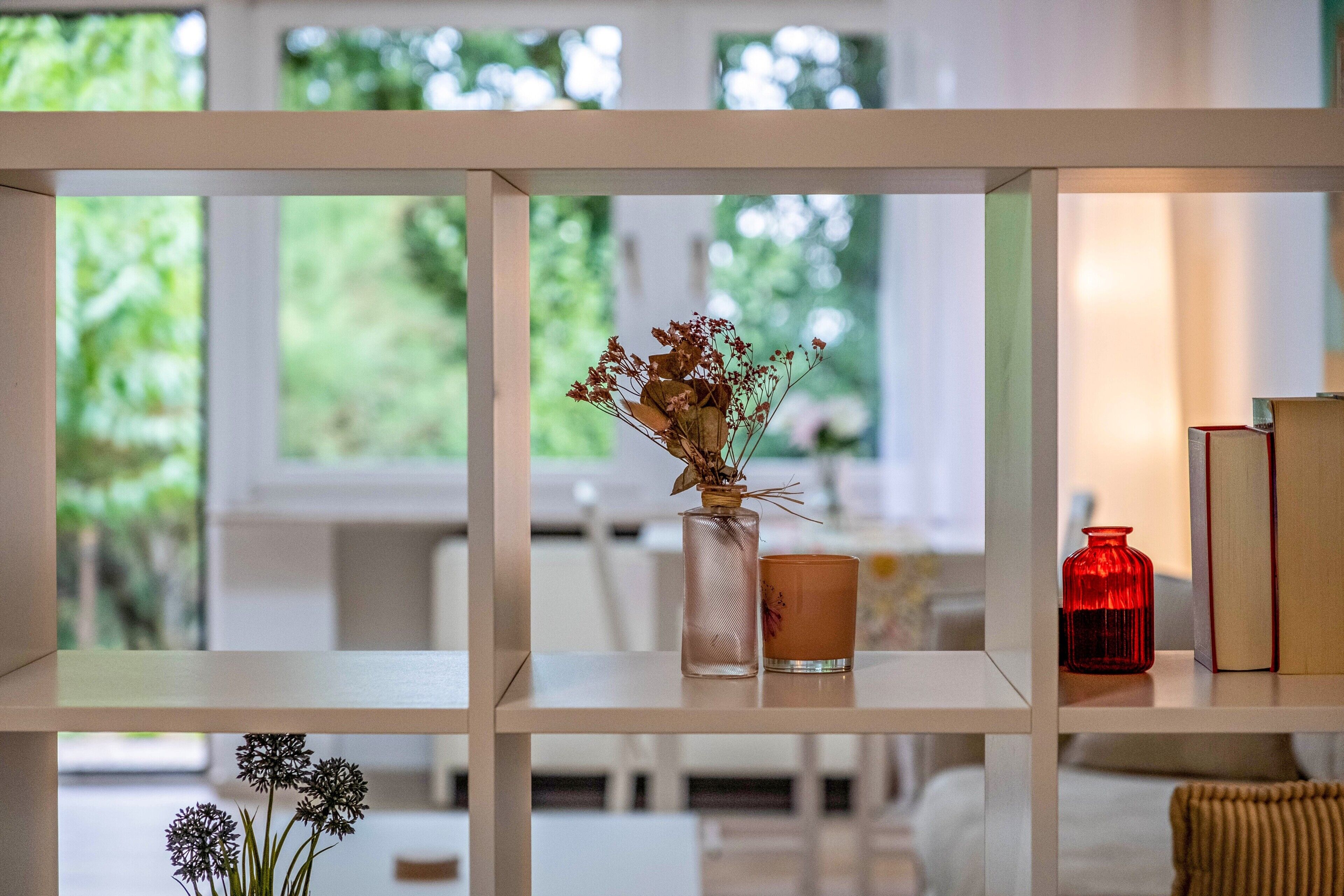 Ein Bücherregal mit einer Vase getrockneter Blumen, einer Kerze und einer roten Flasche.