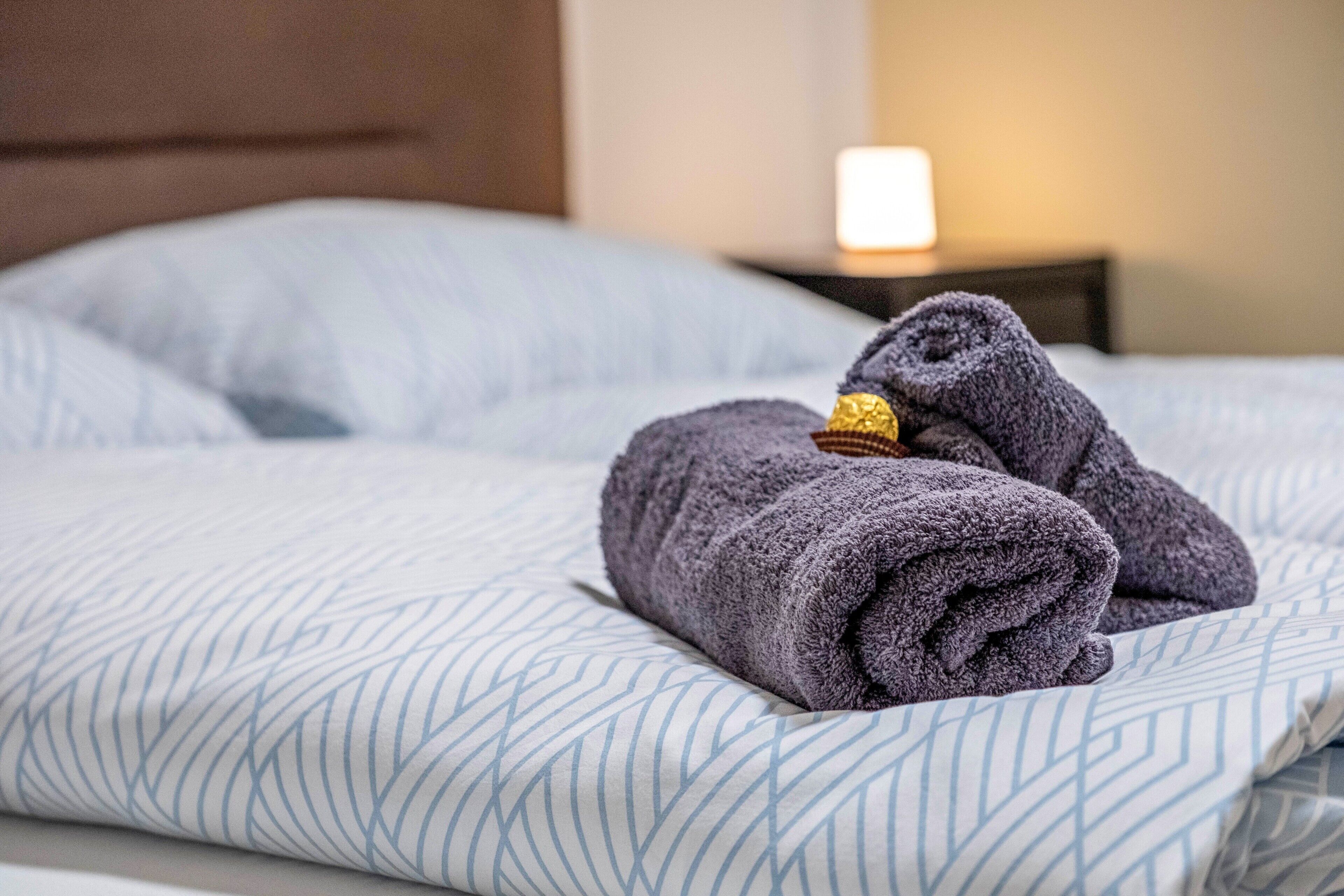 A neatly made bed with a patterned bedspread and two folded towels with a gold tag on a bedside table.