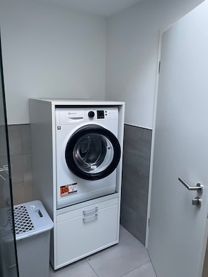 Een compacte, witte wandwasmachine met bedieningspaneel en een klein opbergvakje, geplaatst in een hoek van een kamer met betegelde vloer.