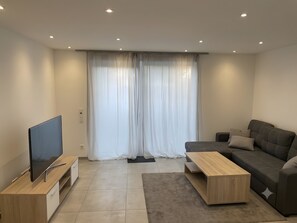 Een moderne woonkamer met een flatscreen televisie op een houten standaard, een grijze bank met kussens, een houten salontafel en een groot raam met dunne gordijnen.