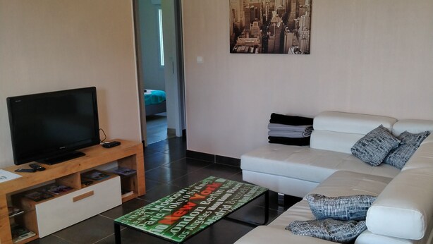 Un salon moderne avec un téléviseur à écran plat posé sur un meuble en bois, un canapé blanc agrémenté de coussins à motifs, une table basse ornée d’une image représentant un paysage urbain et, au mur, un tableau encadré représentant également un paysage urbain.