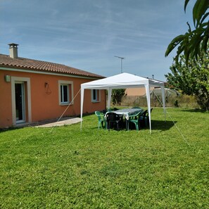 A house with a white tent set up on a grassy lawn, a table with chairs, and a tree in the background.