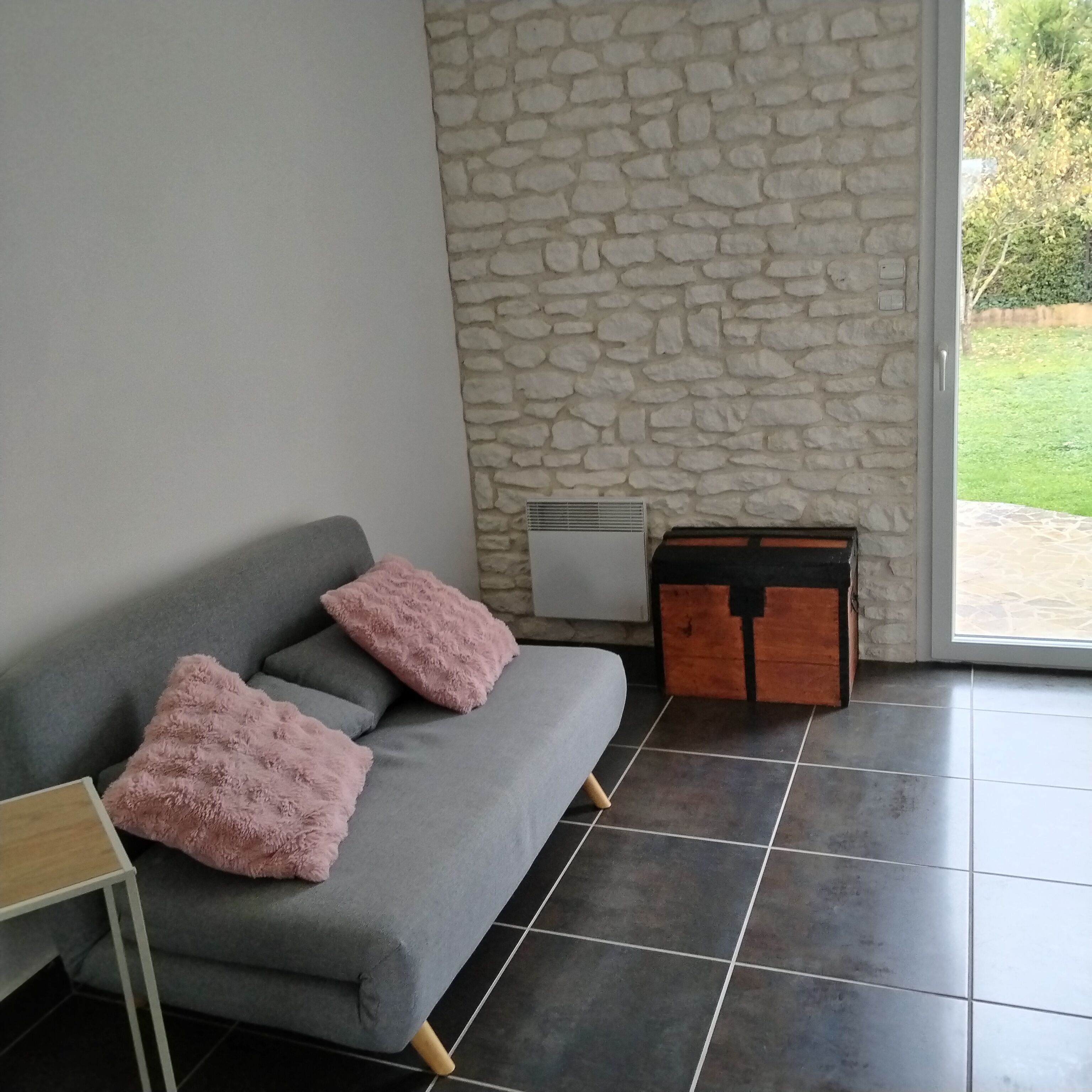 A modern living room with a grey sofa, two pink cushions, a wooden cabinet, and a stone wall.