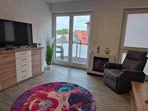 Ein modernes Wohnzimmer mit einem Flachbildfernseher, einem Ledersessel, einem farbenfrohen Teppich und einer Glastür, die auf einen Balkon führt.