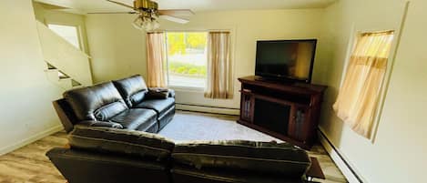 Una sala de estar con un sofá, un televisor sobre un mueble bajo, un ventilador de techo y una ventana con cortinas.