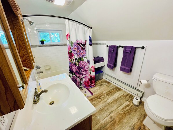 Un baño con lavamanos blanco, ducha con cortina floral y un inodoro con toallas púrpuras.