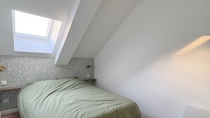 Une pièce avec un plafond en pente, une lucarne, un lit recouvert d’une couverture verte, un plancher en bois et une petite table avec une lampe.