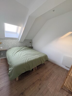 A room with a sloped ceiling, a skylight, a bed with a green bedspread, a wooden floor, and a small table with a lamp. - Vacation Rental in the Heart of the Owner's Stable (Écoust-Saint-Mein)