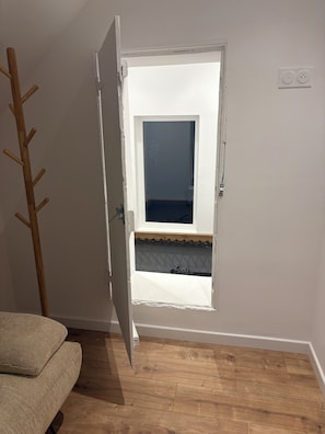 A room with a wooden floor, a white door, and a window with a view of a balcony. - Vacation Rental in the Heart of the Owner's Stable (Écoust-Saint-Mein)