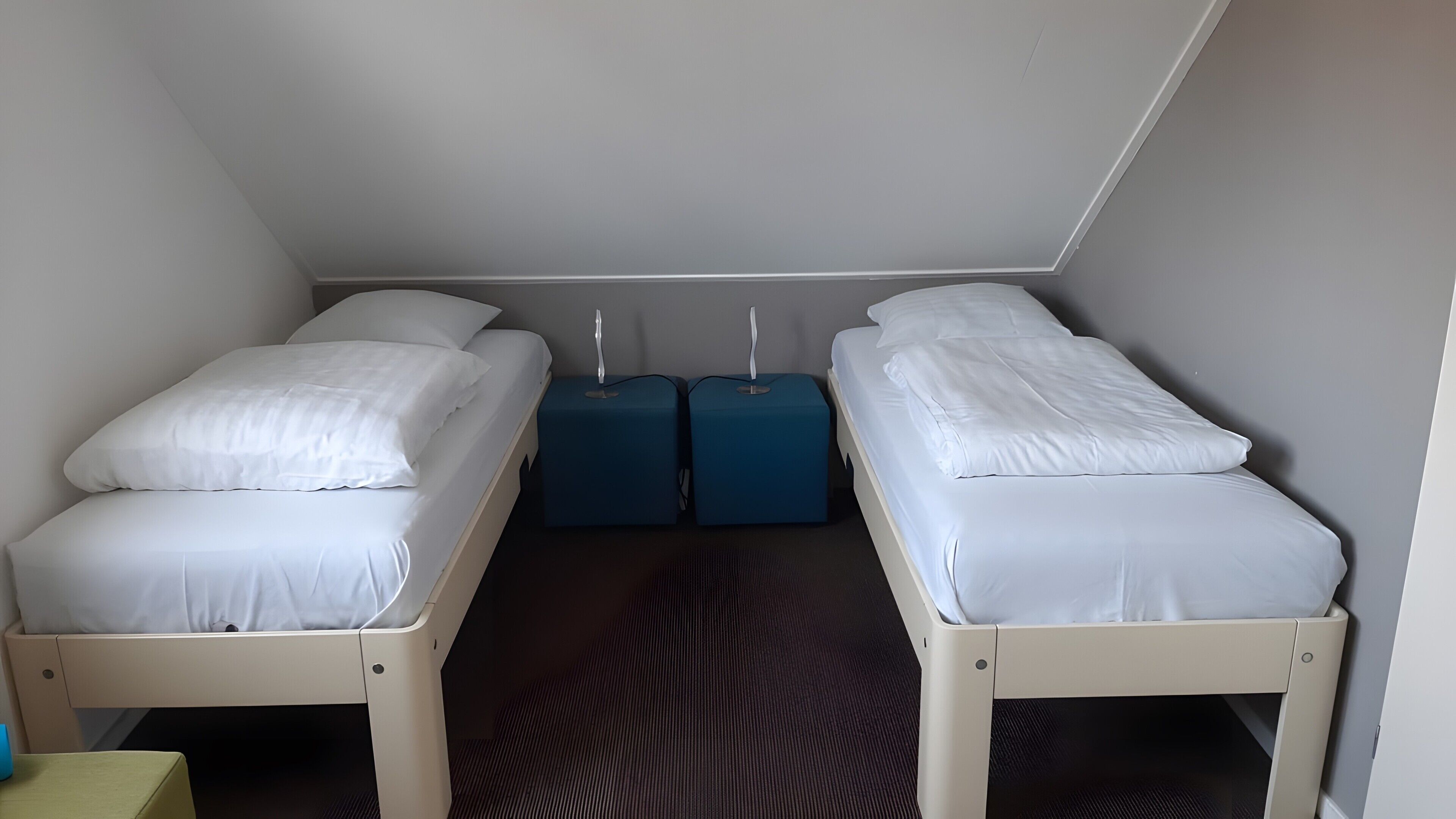 A small, compact room with two single beds, each with white bedding. There are two blue bedside tables with lamps. The room has a sloped ceiling and a patterned carpet.