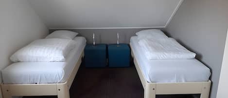 A small, compact room with two single beds, each with white bedding. There are two blue bedside tables with lamps. The room has a sloped ceiling and a patterned carpet.