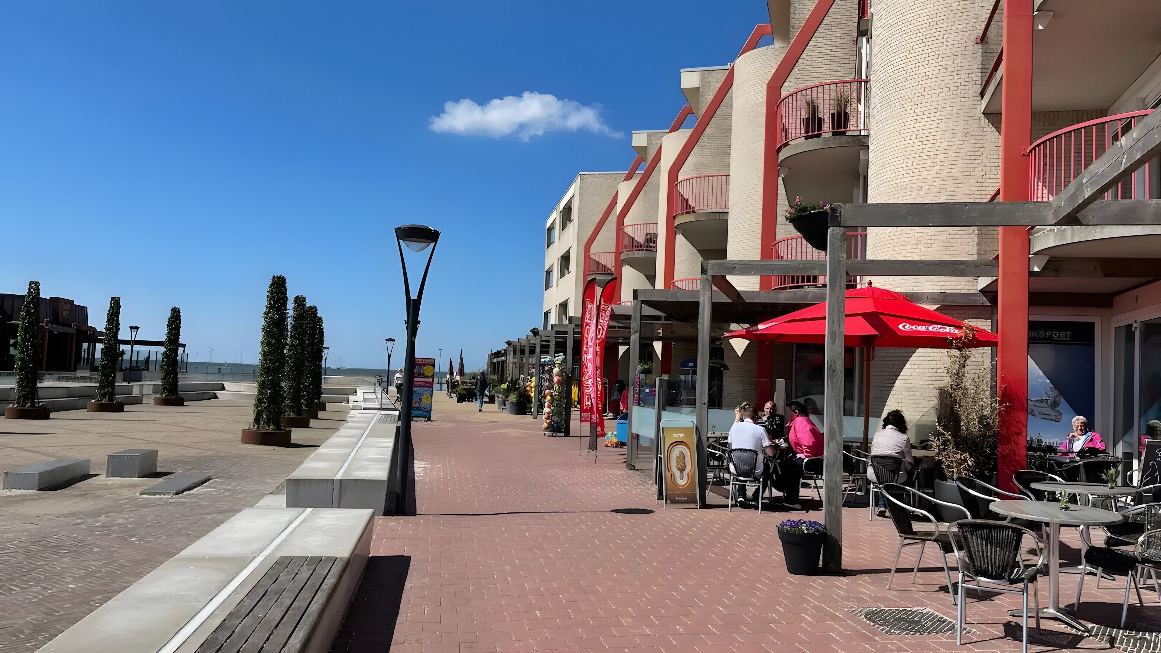 Eine Uferpromenade mit Außenbestuhlung, blauem Himmel und Gebäuden mit Balkonen.