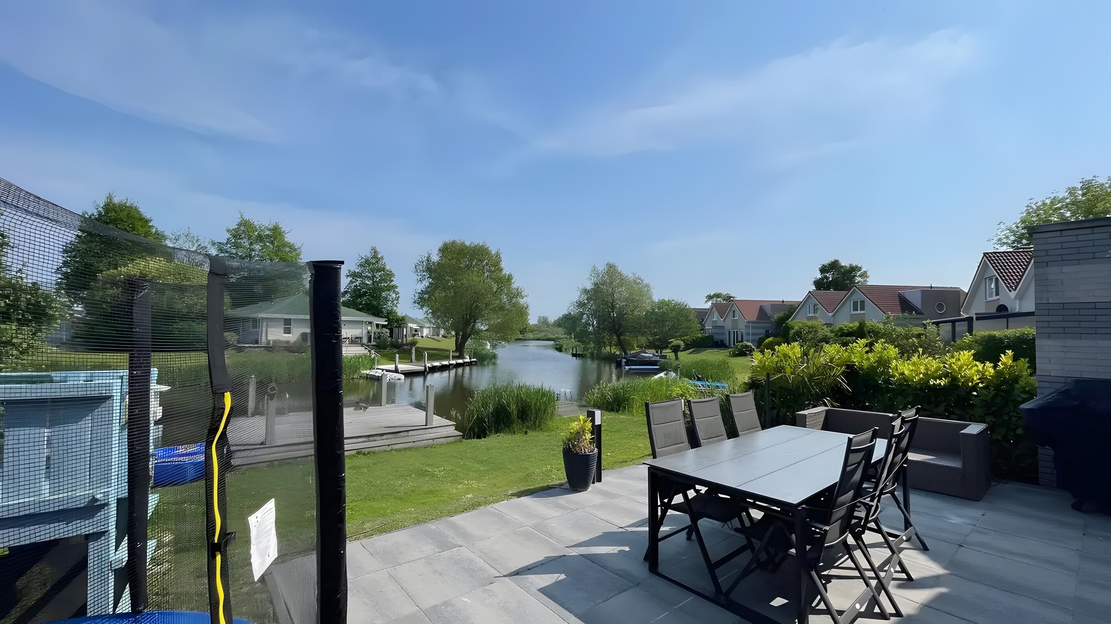 Un patio exterior con una mesa y sillas, vista a un río y casas residenciales al fondo.