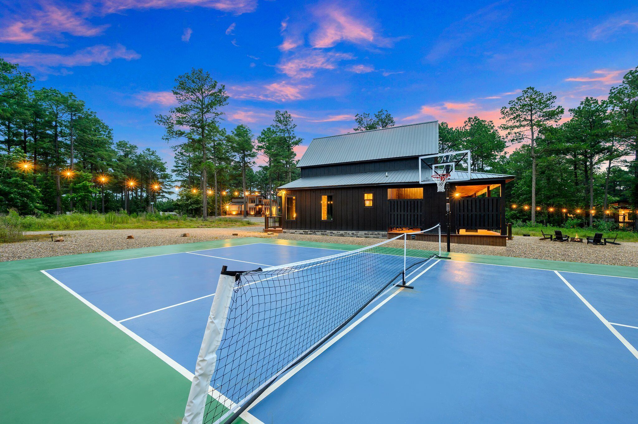 A tennis court with a net in the center, surrounded by a building with a basketball hoop, and trees in the background.