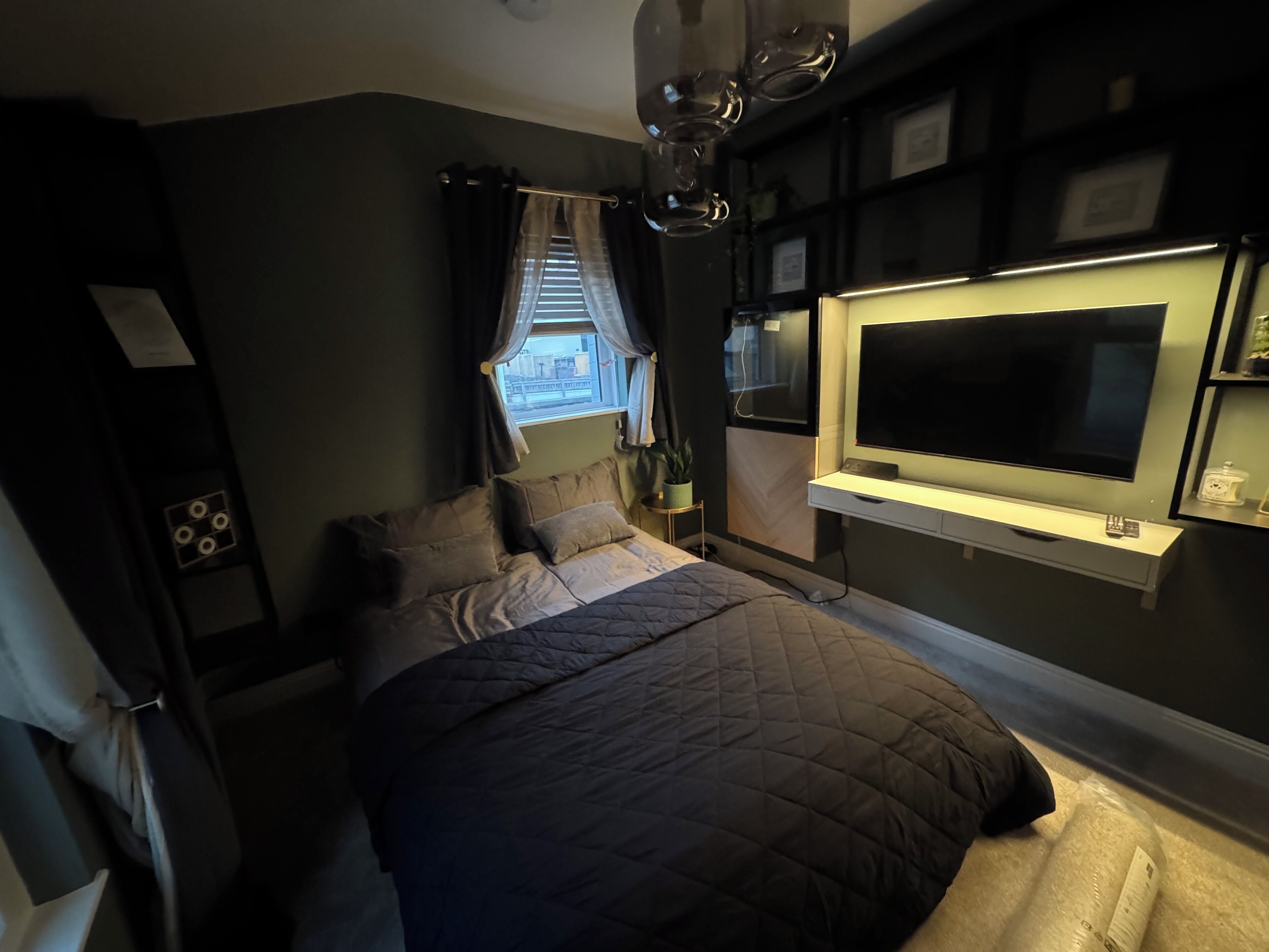 Une chambre à coucher avec un lit, un téléviseur fixé au mur, une fenêtre avec des rideaux et une table de chevet.