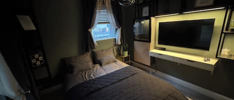 A bedroom with a bed, a TV mounted on the wall, a window with curtains, and a bedside table.