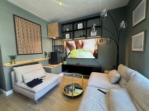 A modern living room with a grey sofa, a small coffee table, a flat-screen TV mounted on the wall, and a bookshelf with decorative items. - Boho Rooms (Bristol)