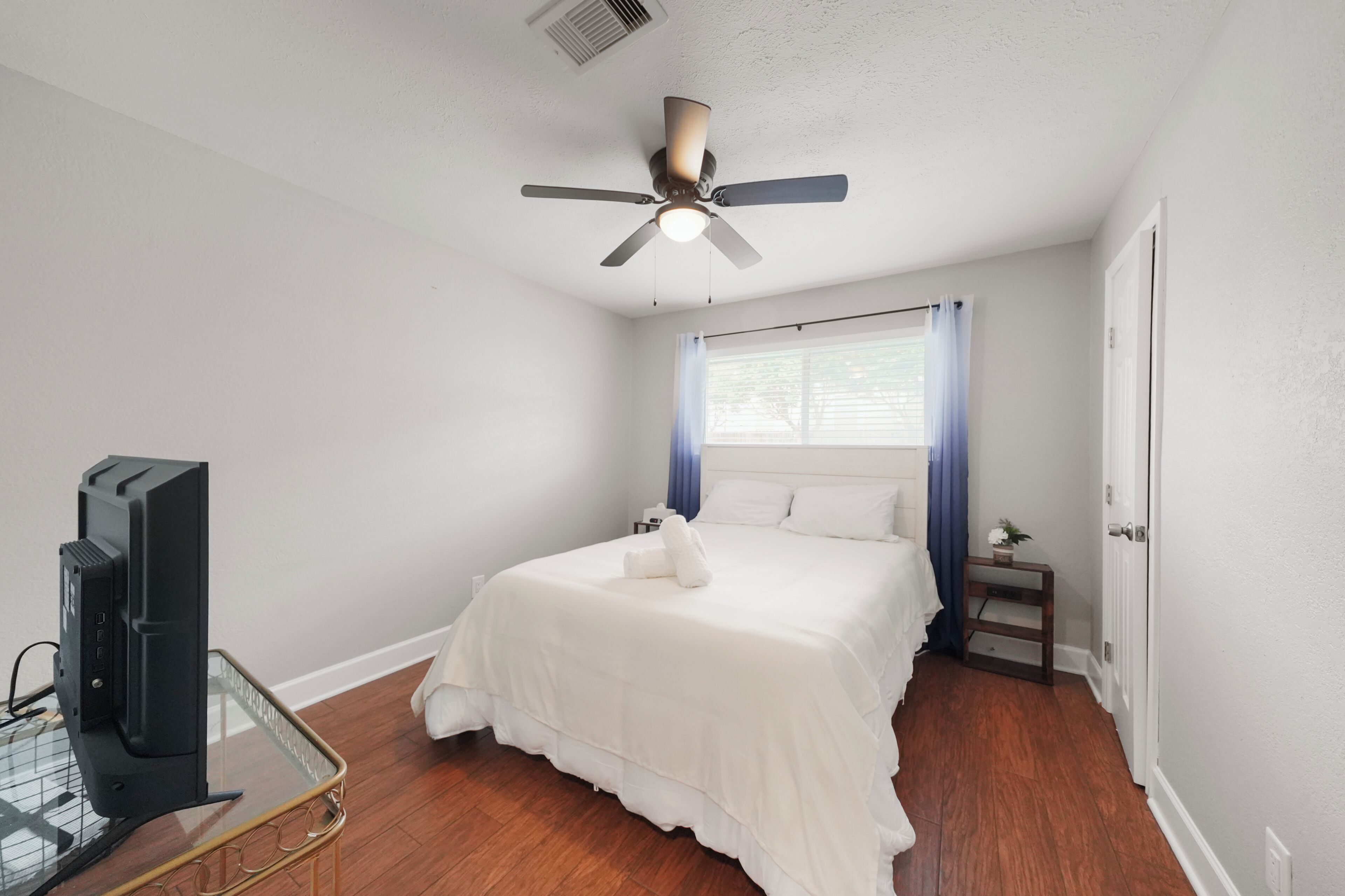 Une chambre à coucher comprenant un lit, un ventilateur de plafond, une fenêtre avec des rideaux, une table de chevet et un téléviseur.