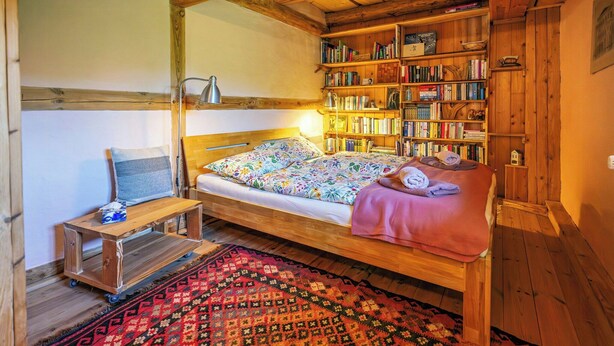 Una habitación acogedora con una cama de madera, una mesita pequeña, una estantería y una alfombra estampada.