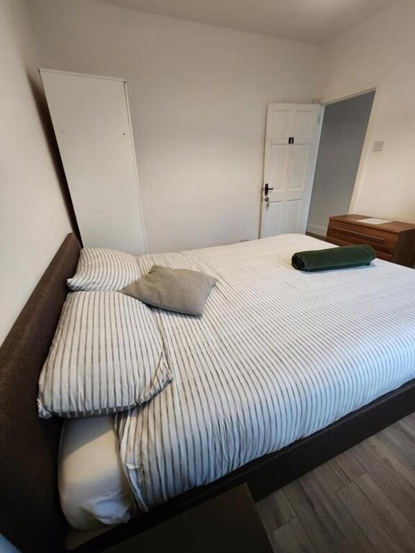 A single-bed room with a wooden headboard, a small bedside table, a wardrobe, and a door leading to another room.