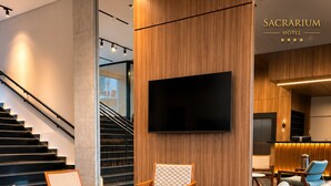 Un moderno vestíbulo de hotel con una escalera, un televisor de pantalla plana montado en una pared de madera y una zona de estar con sillas de madera y una mesa redonda.