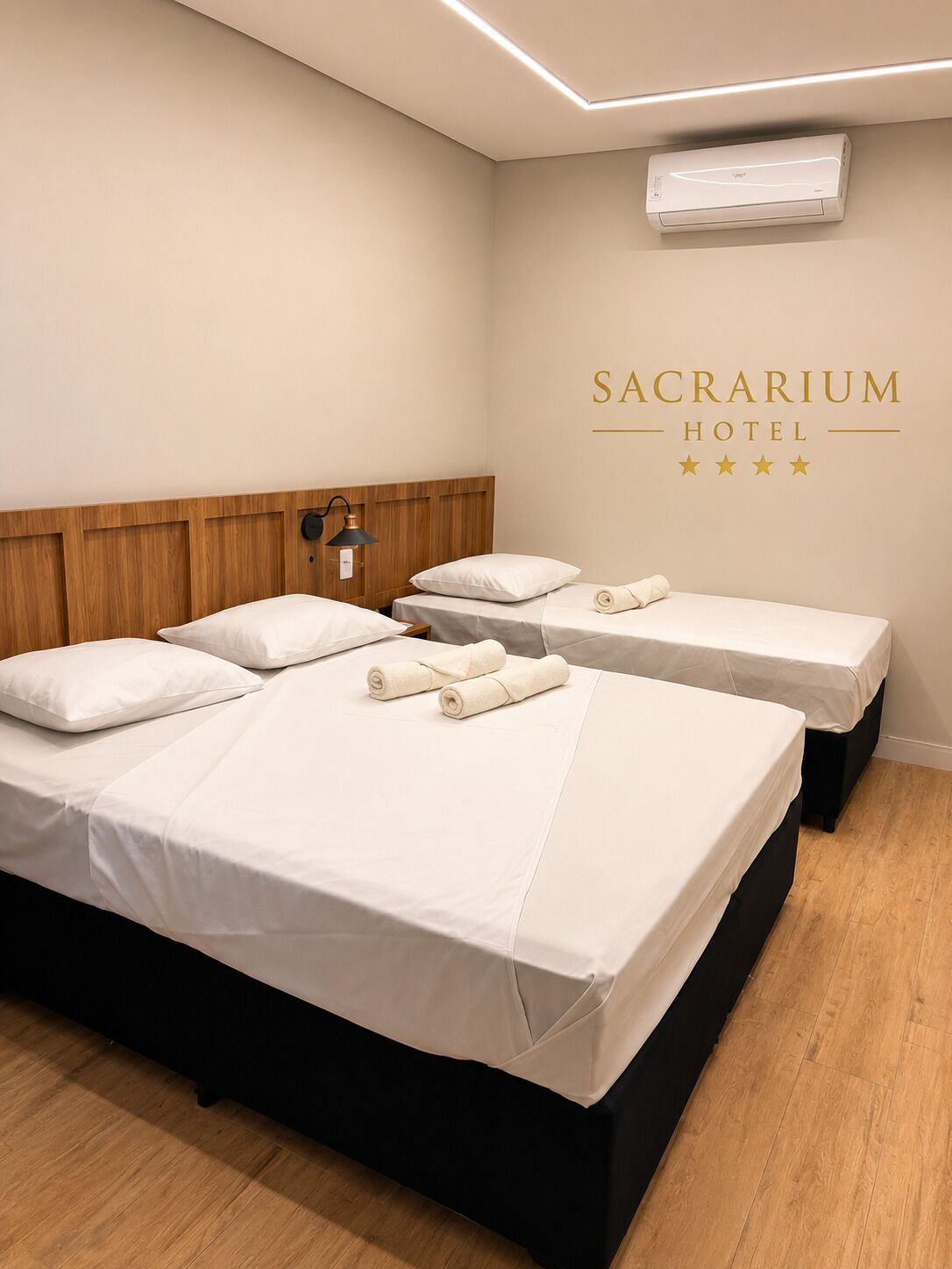 Habitación de hotel con dos camas, cabecera de madera, unidad de aire acondicionado y un letrero en la pared que dice 'SACRARIUM HOTEL'.
