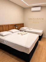 Quarto de hotel com duas camas, uma cabeceira de madeira, um aparelho de ar condicionado e uma placa na parede que diz 'SACRARIUM HOTEL'.
