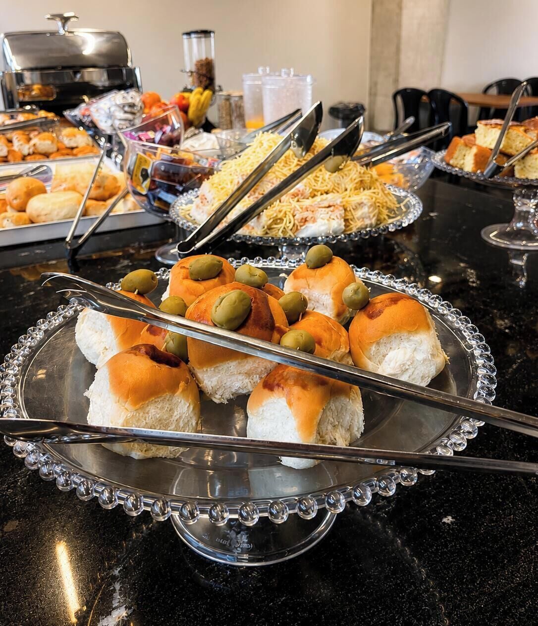 Une table de buffet proposant divers plats, notamment des petits pains aux olives, et un barbecue en arrière-plan.