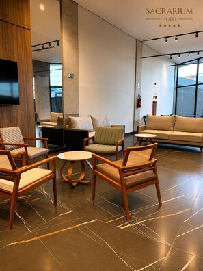 Vestíbulo de hotel con diseño moderno, que incluye un sofá grande, varios sillones, una mesita redonda y un televisor de pantalla plana montado en la pared.