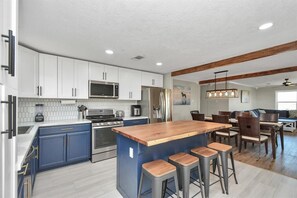 A modern kitchen with white cabinets, a central island with stools, a dining area with wooden chairs, and a ceiling with exposed beams. - Holds 8 · Pool · 3BR/2BA Upper Duplex Peacock Horse Beach Nearby Out City Life (Galveston)