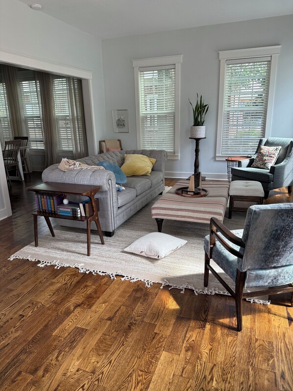 A living room with a grey sofa, a striped ottoman, a wooden coffee table, and a blue armchair. There are two windows with blinds, a floor lamp, and a bookshelf with books.